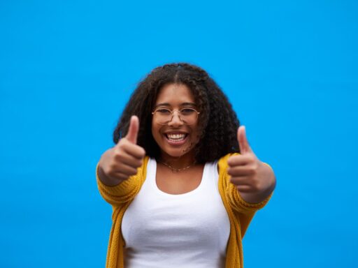 Person in yellow cardigan and glasses giving enthusiastic thumbs up against blue backdrop.